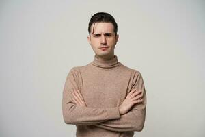 Portrait of handsome young brunette male in trendy wear wearing stylish hairstyle, folding hands on his chest while posing over white background, looking seriously to camera and frowning eyebrows photo