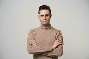 Severe young dark haired man in beige roll-neck sweater standing over white background with folded hands, looking harshly at camera and frowning his eyebrows photo