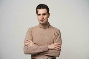 Portrait of young skeptical brunette male with trendy haircut folding hands on his chest and looking at camera with light smile, standing over white background in beige roll-neck sweater photo