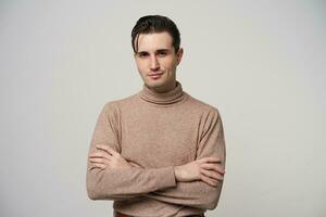 retrato de joven escéptico morena masculino con de moda Corte de pelo plegable manos en su cofre y mirando a cámara con ligero sonrisa, en pie terminado blanco antecedentes en beige cuello vuelto suéter foto