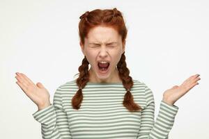 portrait of young ginger female standing over white studio background screaming into camera with closed eyes and raised hands photo