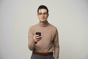 Positive young pretty brunette male hipster with stylish hairstyle holding smartphone and looking at camera with light smile, standing over white background in glasses photo