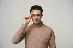 Close-up of handsome young stylish brunette man in beige roll-neck sweater keeping raised hand on his eyewear while looking at camera, isolated over white background photo