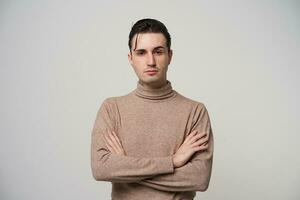 Stylish young pretty dark haired guy with brown eyes keeping hands on his chest and looking severely to camera with raised eyebrow, wearing trendy clothes over white background photo