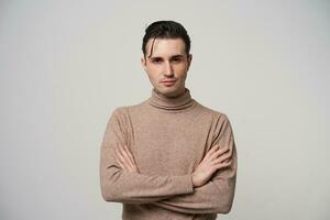 Indoor photo of pretty young brunette male in beige roll-neck sweater crossing hands and looking at camera with folded lips, dressed in trendy clothes while standing over white background