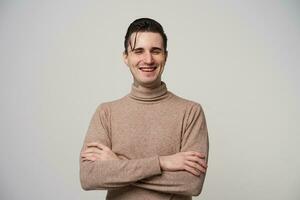 Cheerful beautiful young dark haired man with folded hands laughing happily with closed eyes while posing over white background, wearing beige roll-neck sweater photo