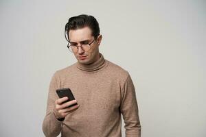 Concentrated young dark haired guy with trendy haircut wearing glasses and stylish clothes while standing over white background, holding mobile phone in hand and looking on screen with folded lips photo