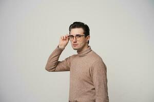 Indoor shot of pretty dark haired guy in glasses wearing stylish hairstyle while posing over white background, looking to camera with folded lips and raised hand photo