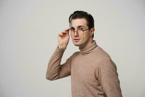 estudio foto de atractivo de moda joven oscuro peludo hombre en lentes posando terminado blanco antecedentes con elevado mano, vestido en beige cuello vuelto suéter