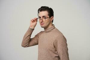 Portrait of handsome brown-eyed young brunette man with trendy haircut holding his eyewear with raised hand and looking at camera with calm face, isolated over white background photo