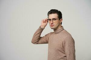 Serious stylish young dark haired man with trendy haircut frowning eyebrows while looking at camera and holding his eyewear with raised hand, standing over white background in fashionable clothes photo