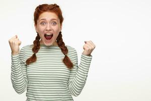 Indoor portrait of young ginger female standing over white background raised her fist with positive facial expression, celebrating a victory photo