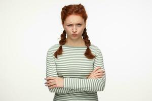 Indoor portrait of young ginger female standing over white background looking into camera with irritated facial expression, keeps her hands crossed photo