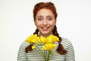 Indoor portrait of young ginger female posing over white background holding bouquet of flowers and smile broadly photo