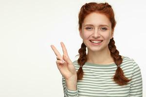 Indoor portrait of young ginger female standing over white background showing v sign into camera photo
