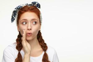 portrait of young ginger female standing over white studio background looks aside with shocked facial expression and whisper a secret photo
