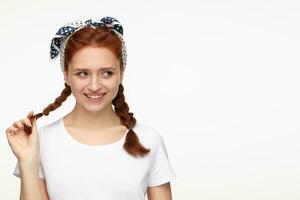 portrait of young ginger female standing over white studio background looks aside at copy space and smiles broadly photo