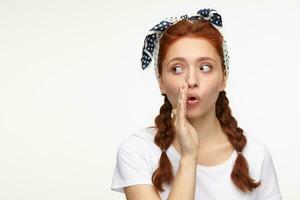 portrait of young ginger female standing over white studio background looks aside with shocked facial expression and whisper a secret photo