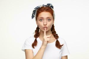 portrait of young ginger female standing over white studio background shows silence gesture with socked facial expression photo