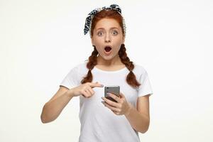 ginger young female points with a finger at phone with shocked facial expression photo