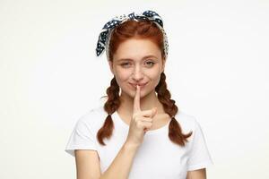 portrait of young ginger female standing over white studio background raised up eyebrow and shows silence gesture while flirting photo