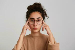 retrato de concentrado pensativo georgiano joven mujer con Rizado pelo usa beige pull-over y lentes conmovedor su templos y pensando aislado terminado blanco antecedentes foto