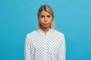 Portrait of upset displeased blonde young woman wears polka dot shirt makes sad face and feels depressed isolated over blue background photo