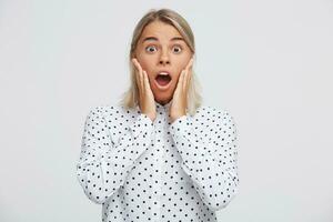 Closeup of amazed astonished blonde young woman with opened mouth wears polka dot shirt feels shocked and surprised and shouting isolated over white background photo