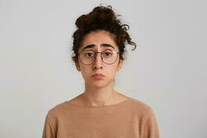 de cerca de triste Deprimido georgiano joven mujer con Rizado pelo usa beige pull-over y gafas siente trastornado y decepcionado aislado terminado blanco antecedentes foto