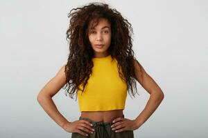 Indoor photo of young attractive long haired curly dark skinned lady holding hands on her waist while looking defiantly at camera, isolated over white background