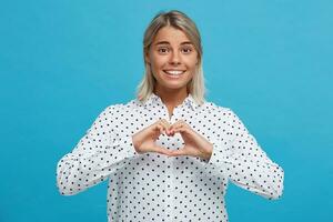 retrato de contento encantador rubia joven mujer usa polca punto camisa sonriente y demostración corazón forma por manos aislado terminado azul antecedentes foto