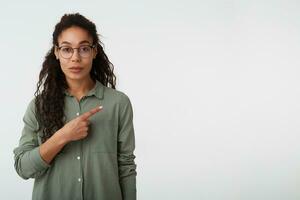 Indoor shot of young attractive curly brunette dark skinned lady keeping her lips folded while pointing aside with forefinger, isolated over white background photo