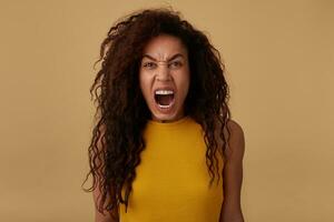 Portrait of angry brown haired curly brunette lady screaming madly while looking at camera and keeping her hands down while posing over beige background photo