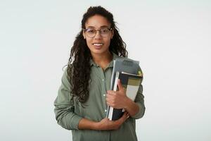 Cheerful young lovely brown haired curly female with dark skin holding books while looking gladly at camera with broad smile, isolated over white background photo