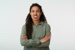 Studio shot of young stressed dark haired curly female with casual hairstyle showing her teeth while grimacing face and keeping hands crossed, isolated over white background photo