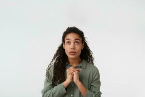 estudio Disparo de joven bonito largo peludo morena dama con oscuro piel plegable elevado manos mientras mirando Ojalá hacia arriba, posando terminado blanco antecedentes foto