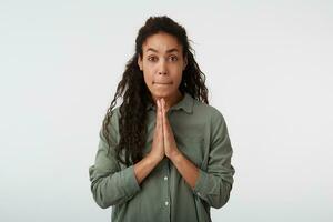 Bewildered young attractive long haired curly dark skinned woman with natural makeup raising hands in praying gesture and biting worringly her lips while posing over white background photo