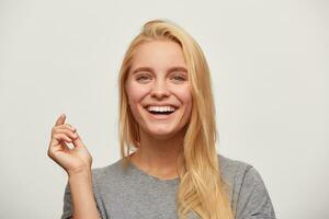 Close up of laughing beautiful blonde young woman, sole of company, spends time with close friends, feels happy, one hand up, toothy smile, wears grey t-shirt, isolated over white background photo