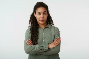 Confused young attractive dark haired curly female keeping her hands crossed and raising surprisedly eyebrow while looking at camera, standing over white background photo