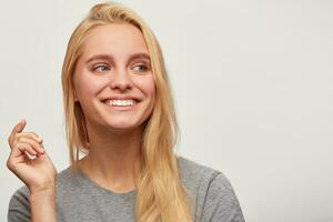 retrato de riendo bonito rubia joven mujer, gasta hora con cerca amigos compañía, siente feliz, uno mano arriba, mirando aparte, con dientes sonrisa, usa gris camiseta, aislado terminado blanco antecedentes foto