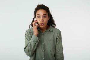 Indoor photo of excited young dark haired curly female with natural makeup rounding surprisedly her brown eyes while having phone talk, isolated over white background