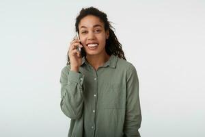 Studio shot of cheerful young lovely dark haired curly dark skinned woman smiling pleasantly while having phone talk, standing over white background in green shirt photo