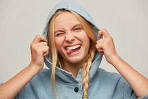 Close up of nice joking beautiful blonde young woman with braid, hands keep hood, feels happy, having fun, shows tongue, wears oversize jeans denim coat isolated over white background photo