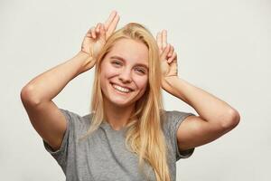 cerca arriba de bonito bromas hermosa rubia joven mujer, siente feliz, hace cuernos en su cabeza con dedos, teniendo divertido, con dientes sonrisa, usa gris camiseta, aislado terminado blanco antecedentes foto