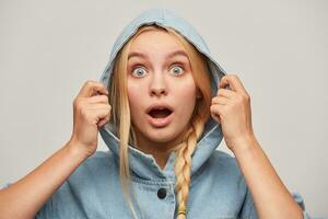 Portrait of amazed beautiful blonde young woman with braid, hands keep hood, looks open-eyed, wears oversize denim coat very surprised or shocked, over white background photo