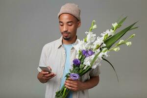 retrato de joven alegre atractivo chico en gris sombrero, sostiene un ramo de flores en su manos, mira a el teléfono inteligente y charlando, soportes terminado gris fondo foto