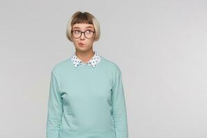 Closeup of thoughtful pensive young woman wears blue sweatshirt and spectacles looks pensive, thinking and looks to the side isolated over white background photo