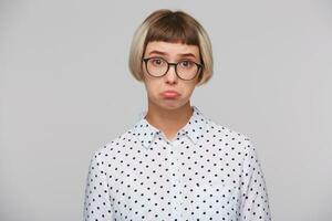 Closeup of sad upset blonde young woman wears polka dot shirt and spectacles curving lips and feels depressed isolated over white background photo