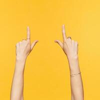 Studio photo of pretty woman raised hands with ornamentals showing upwards with index fingers while being isolated over yellow background. Signs and gestures concept