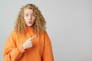 Horizontal shot of shocked young female, expresses great surprisment. Surprised girl keeps mouth open, being stunned, points to the right side with index finger on copy space, over white background photo